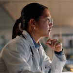 Person in a lab coat and glasses, seated in profile view with a thoughtful expression.