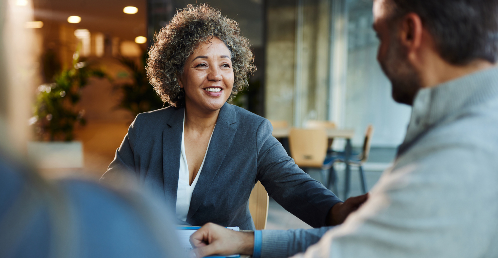 A smiling woman in a dark blazer conversing with a man in a modern office setting.