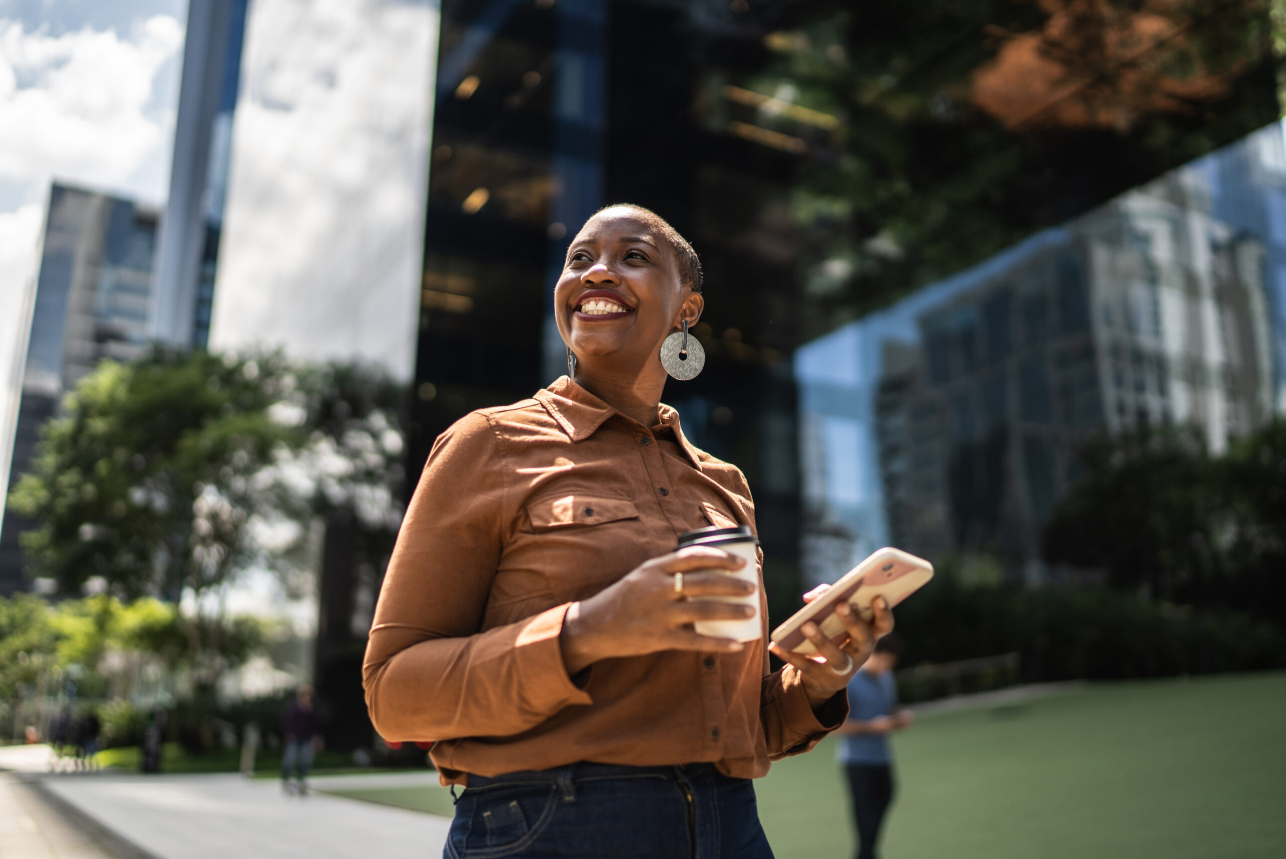 A person standing outdoors holding a coffee cup and smartphone, smiling and looking into the distance, with modern glass buildings and greenery in the background.