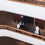 Overhead view of three people in business attire walking in a curved corridor with wooden railings and white walls.