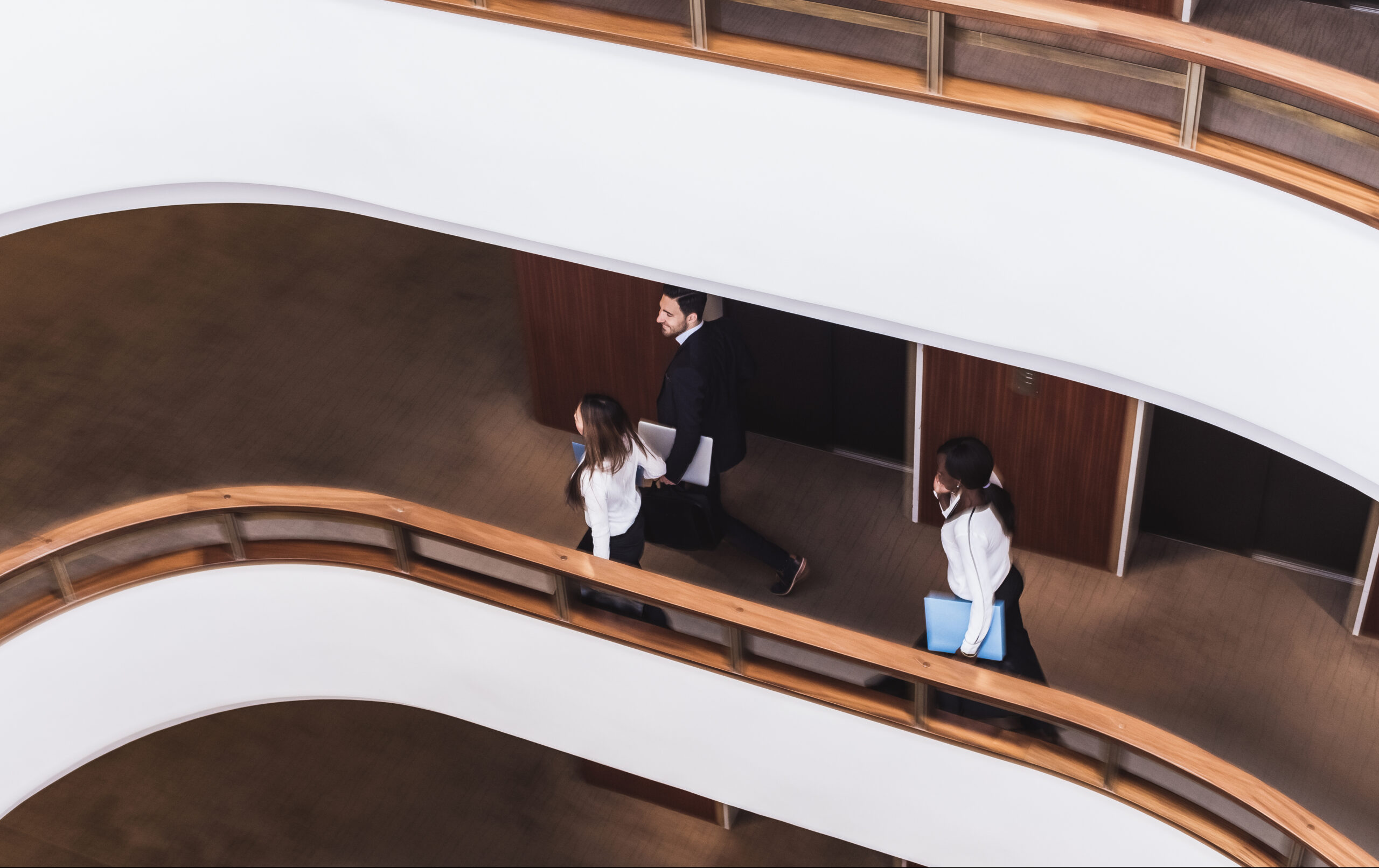 Overhead view of three people in business attire walking in a curved corridor with wooden railings and white walls.