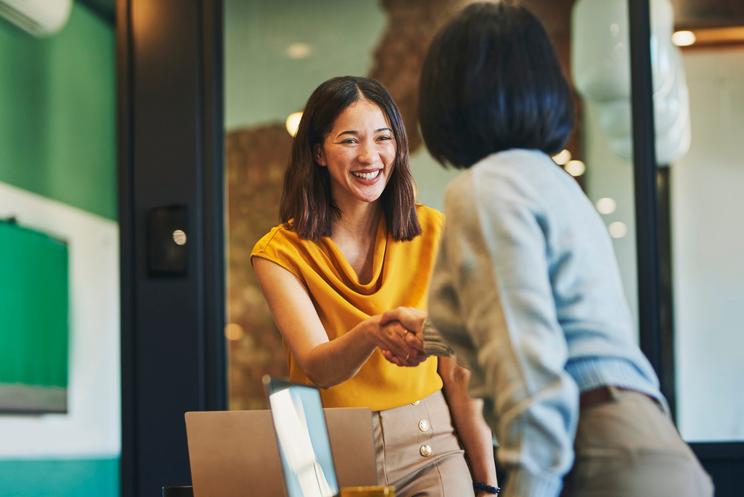 Two women shaking hands in an office setting, one smiling widely.