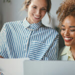 Two women smiling while looking at a laptop screen together.