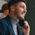 Man in a blue blazer smiling while on a phone call in an office setting.