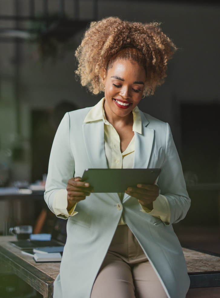 Person smiling at a tablet while seated on a table in a blazer and trousers.