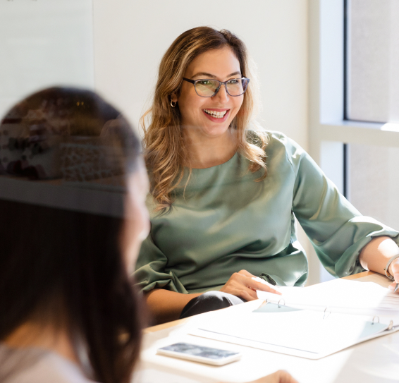 Two people in a bright room talking, with one woman smiling and an open binder on the table.