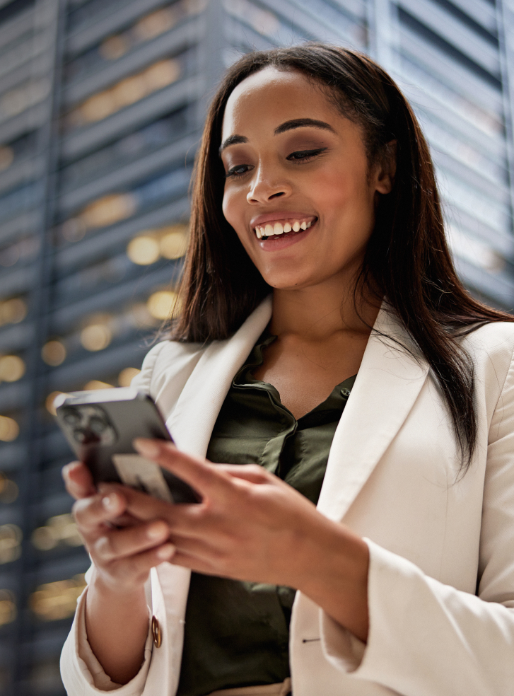 A woman in a white blazer smiles at her smartphone with a skyscraper in the background.