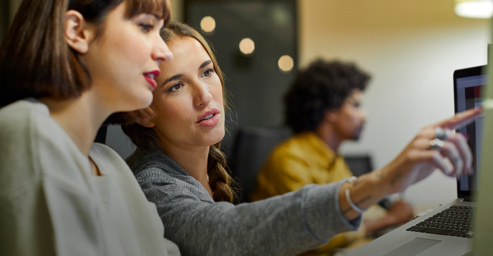 Two women discussing something on a laptop screen; a man works in the background.