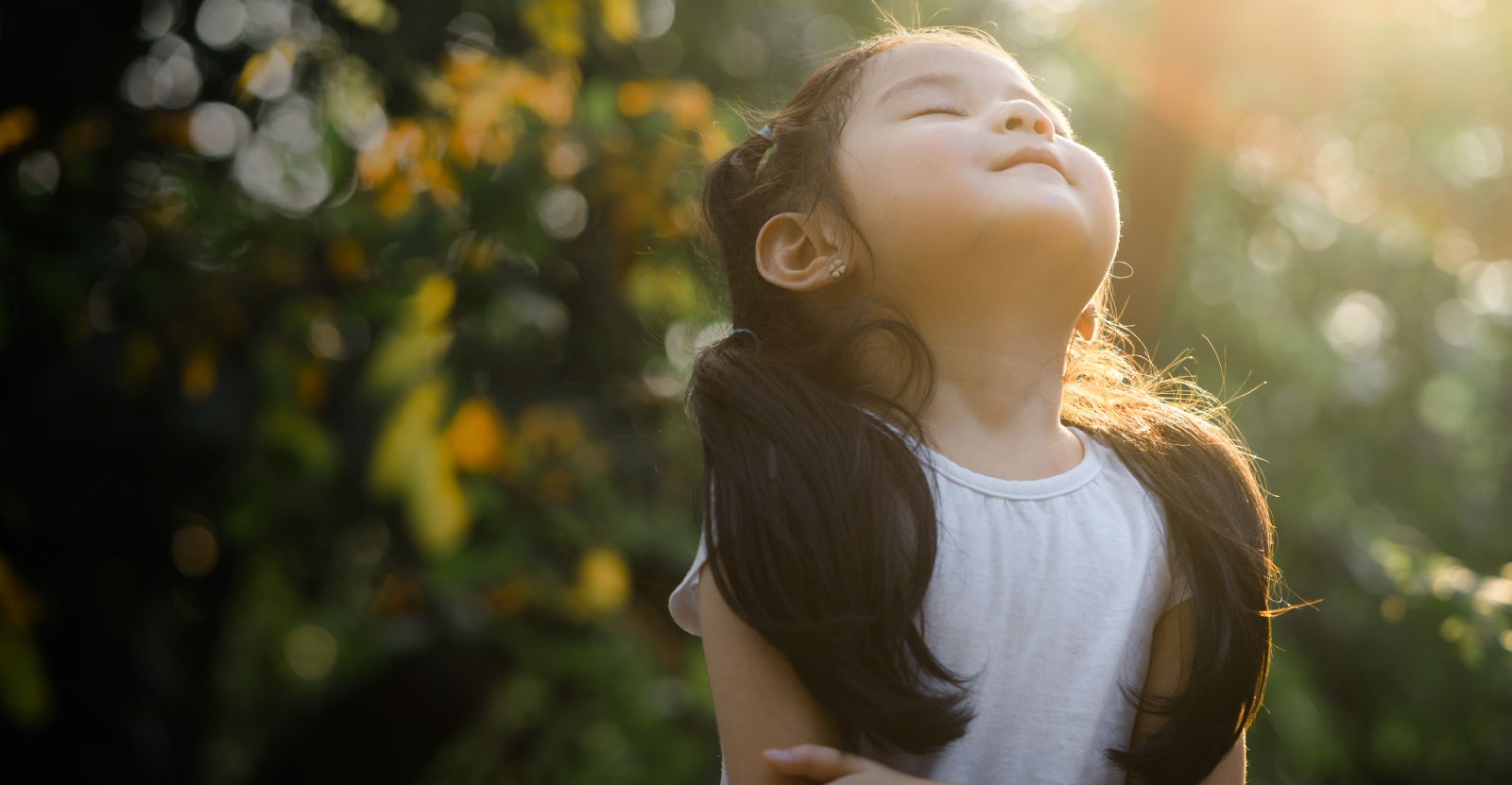 Young girl with pigtails enjoying sunlight, eyes closed, in a leafy outdoor setting.