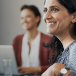 Smiling woman at a meeting table with two blurred colleagues in the background.