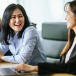 Smiling woman in a light blue shirt at a table with a laptop.