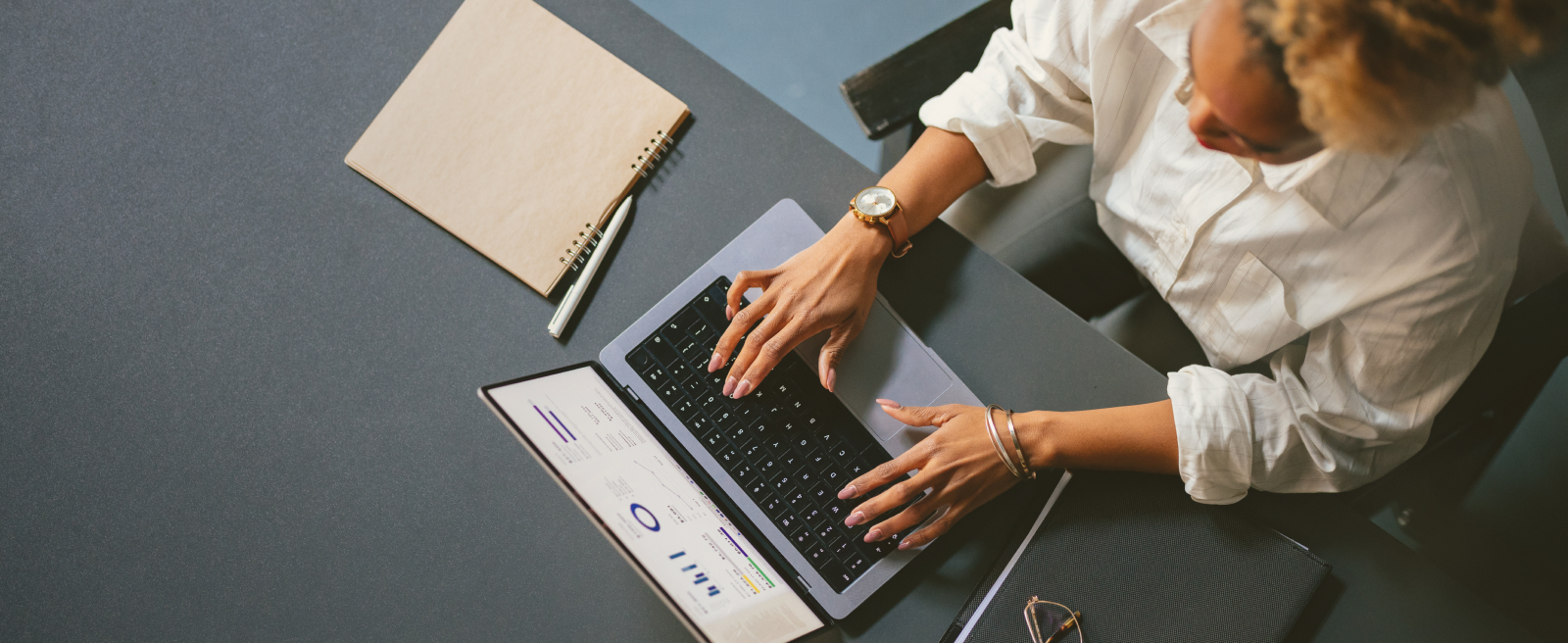 Person typing on a laptop at a desk with a notebook and pen beside them.