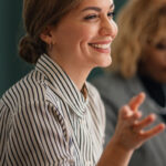 Smiling woman in a striped shirt, sitting at a table, with a blurred person in the background.