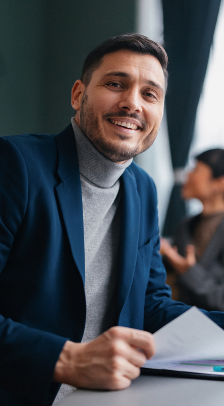 Man in blue blazer and gray turtleneck smiling while holding papers at a desk.