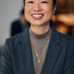 Woman in a gray blazer with arms crossed, smiling in a blurred indoor setting.