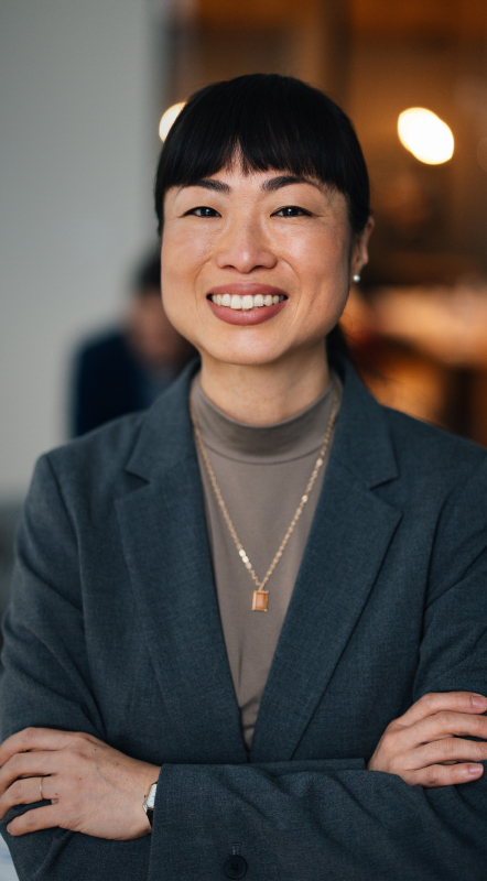 Woman in a gray blazer with arms crossed, smiling in a blurred indoor setting.