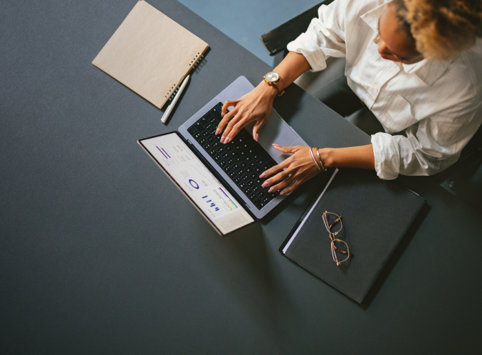 Person typing on a laptop at a dark grey desk with notebooks and glasses.