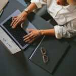 Person typing on a laptop at a table with a notebook and eyeglasses.