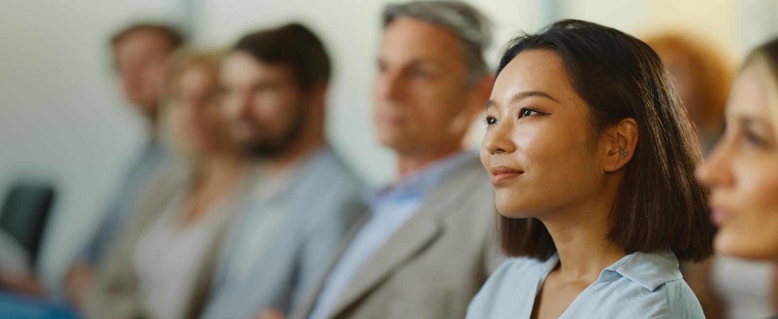 A woman in a light blue shirt listening attentively, with others blurred in the background.