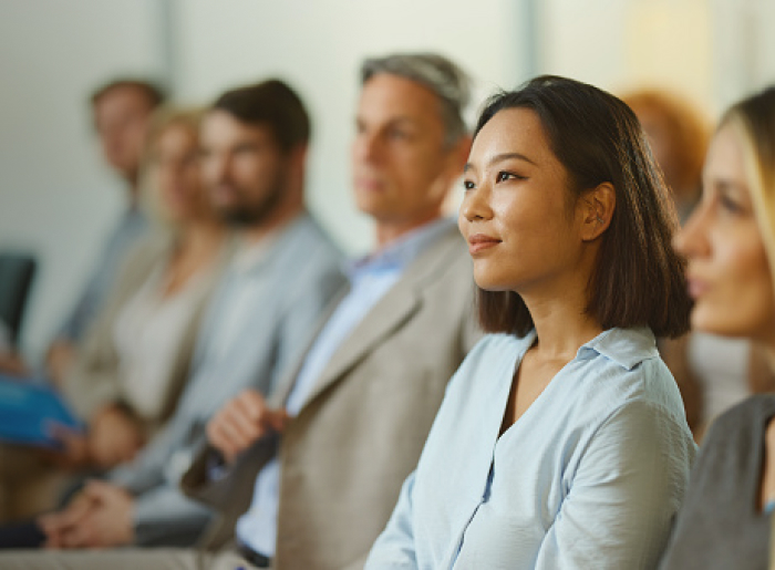 A woman in a light blue shirt listening attentively, with others blurred in the background.