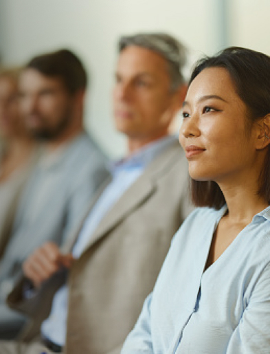 A woman in a light blue shirt listening attentively, with others blurred in the background.