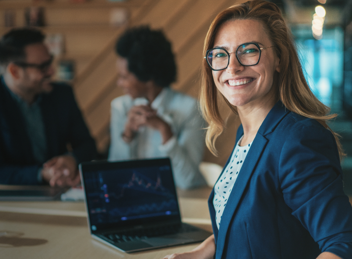 Woman in blue blazer smiling in an office with a laptop showing a graph.