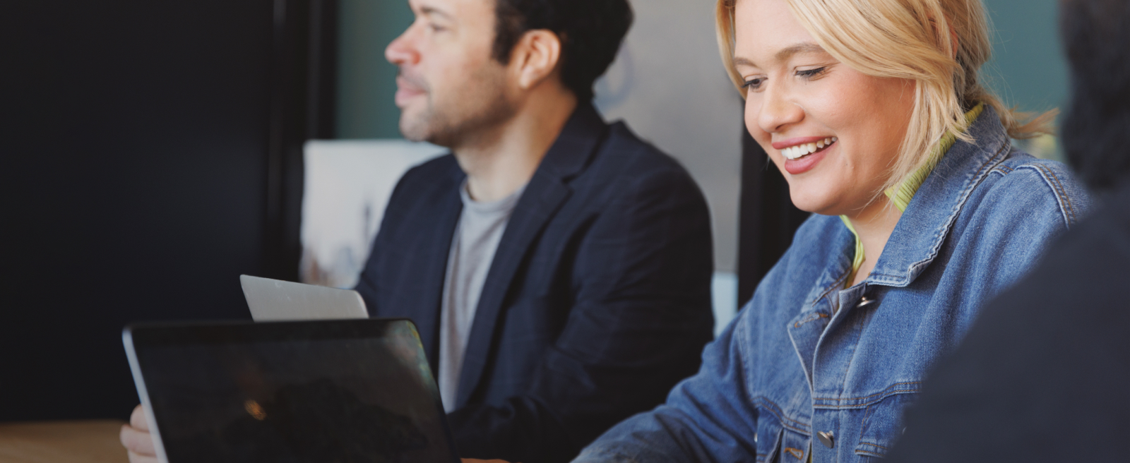 Two people working on laptops, a smiling woman in a denim jacket and a man in a blazer.