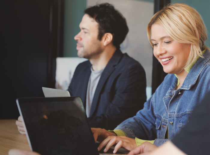 Two people working on laptops, a smiling woman in a denim jacket and a man in a blazer.