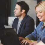 Two people working on laptops at a table. One is a woman smiling, and the other is a man looking to the side.