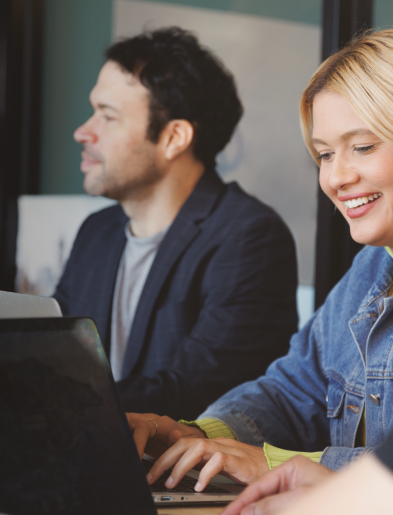 Two people working on laptops, a smiling woman in a denim jacket and a man in a blazer.