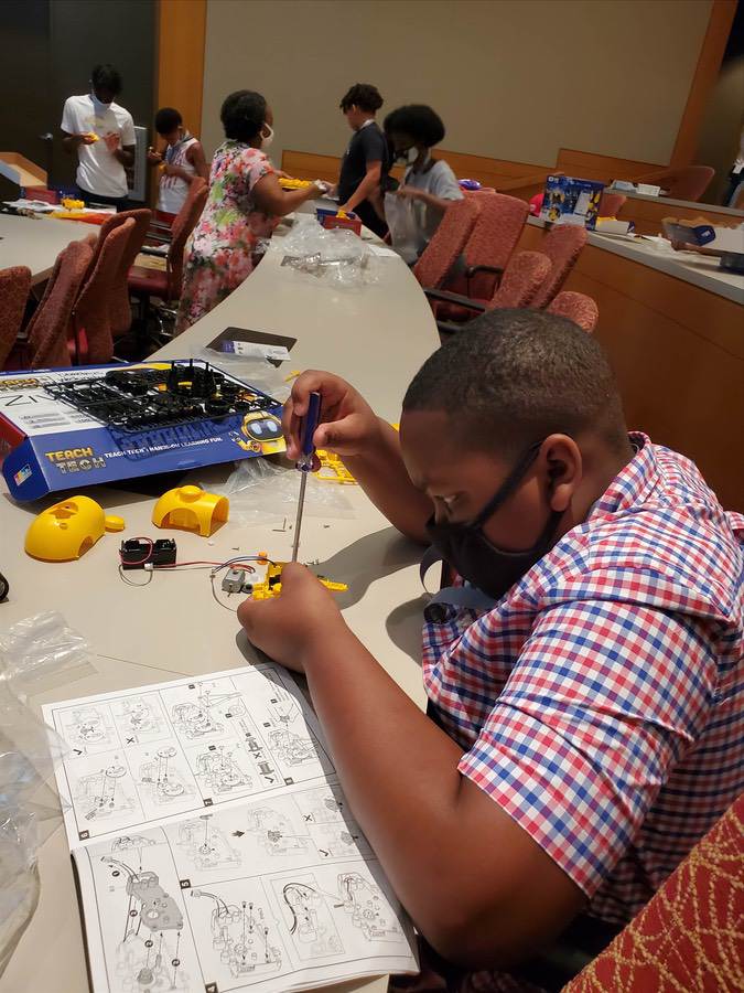 Children assembling parts with a Teach Tech kit in a conference room, one child uses a screwdriver at a table.