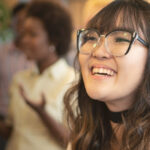 Close-up of a smiling person with glasses, with people conversing in the blurred background.