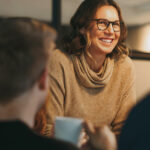 Woman in beige sweater smiling and talking with two people, one holding a white mug.