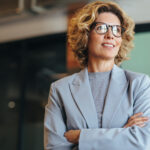 Woman with curly hair wearing glasses and a light blue blazer smiles gently.