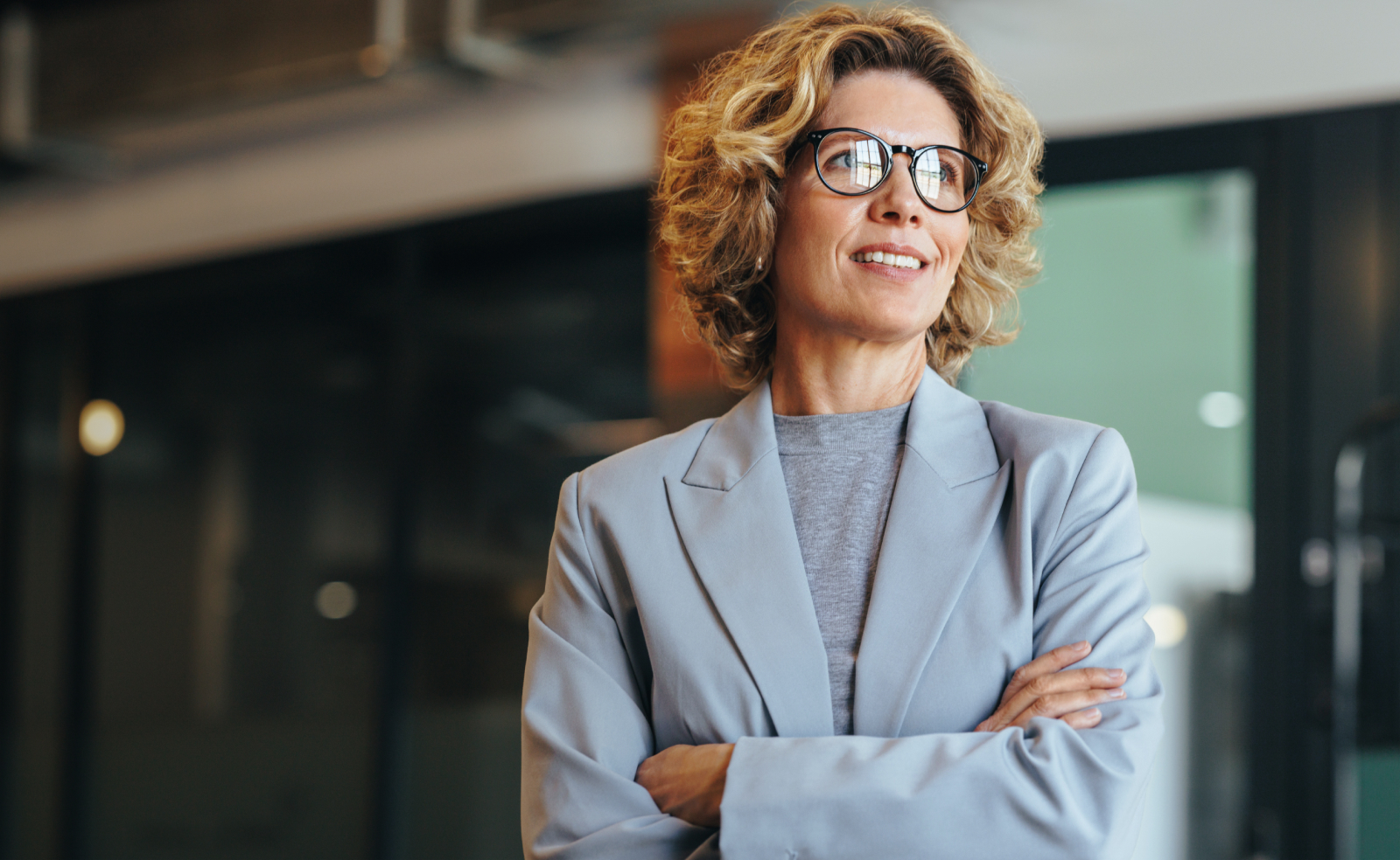 Woman with curly hair wearing glasses and a light blue blazer smiles gently.