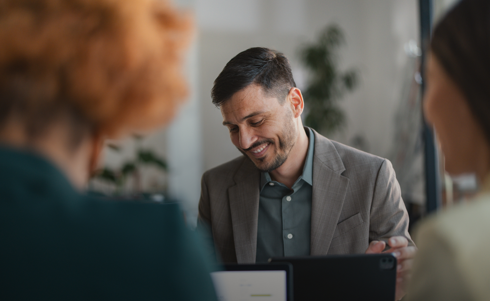 Man in a gray blazer smiling at a tablet in an office setting.