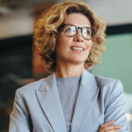 Woman with curly hair wearing glasses and a light blue blazer smiles gently.