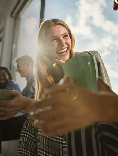 Group of people laughing and holding mugs near a large window with sunlight streaming in.