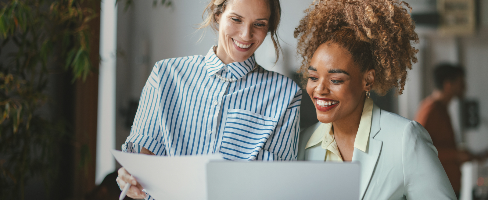 Two women smiling while looking at a laptop screen in an office setting.