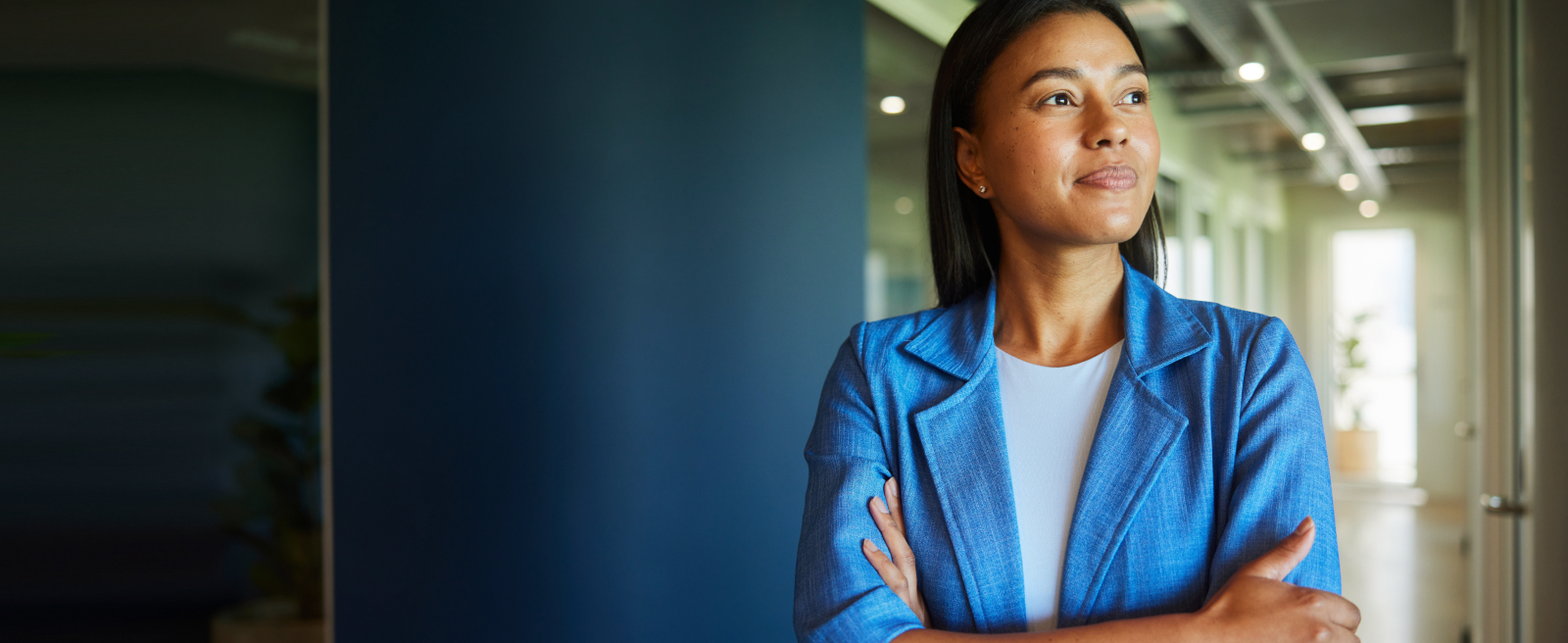 A woman in a blue blazer stands with arms crossed in a modern office hallway, looking to the side.