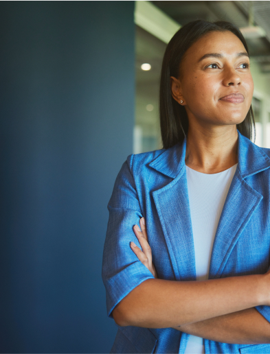 A woman in a blue blazer stands with arms crossed in a modern office hallway, looking to the side.