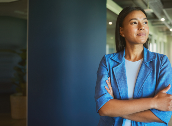 A woman in a blue blazer stands with arms crossed in a modern office hallway, looking to the side.