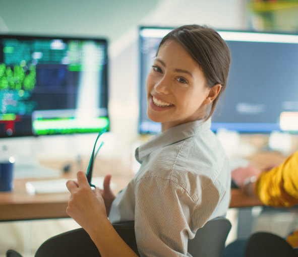 A woman at a desk with multiple monitors turns around in her chair and smilles over her shoulder