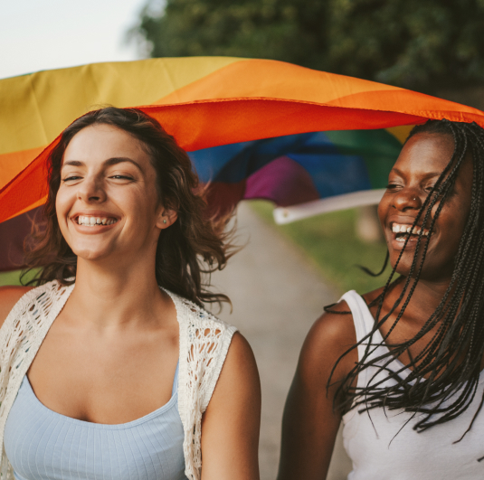 Two people smiling, holding a rainbow flag over their heads outdoors.