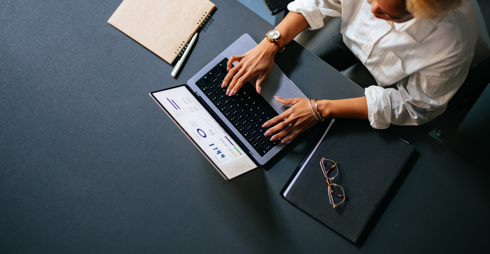 Overhead view of a person typing on a laptop at a grey desk with notebooks, glasses, and a pen.