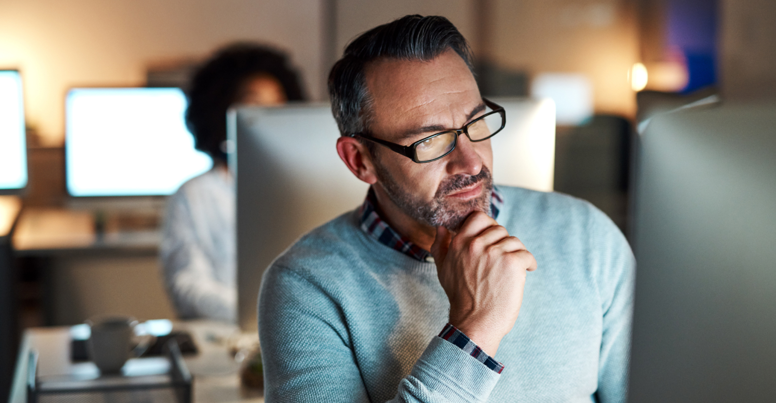 Man in a light blue sweater and glasses working intently at a computer in an office.