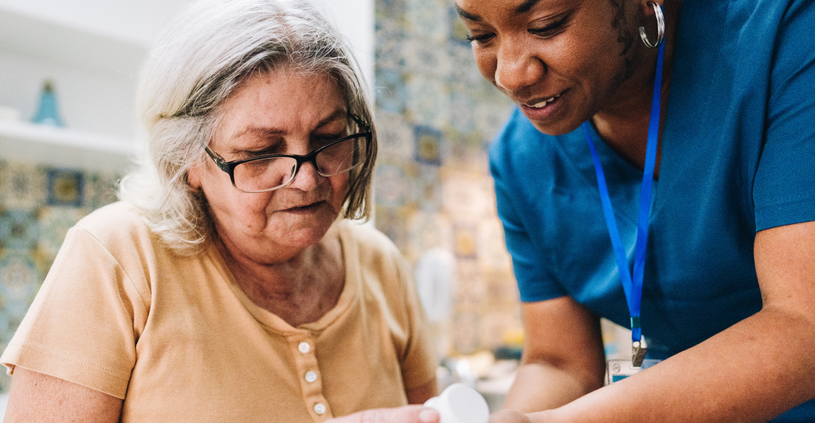 An elderly person in a yellow shirt is assisted by a person in blue scrubs.