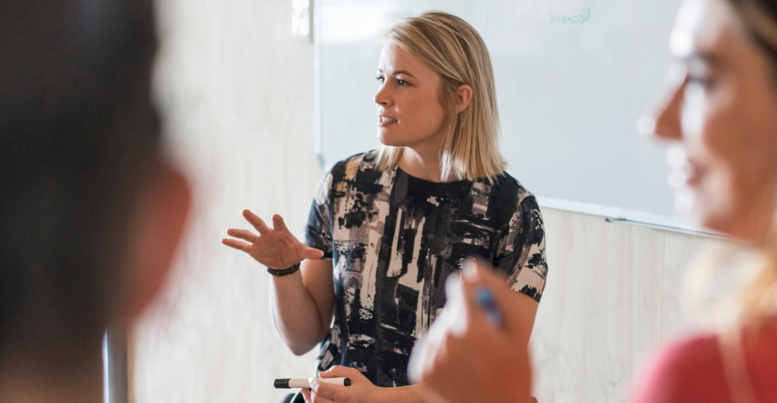 Woman presenting in front of a whiteboard while holding a marker, with people in foreground.