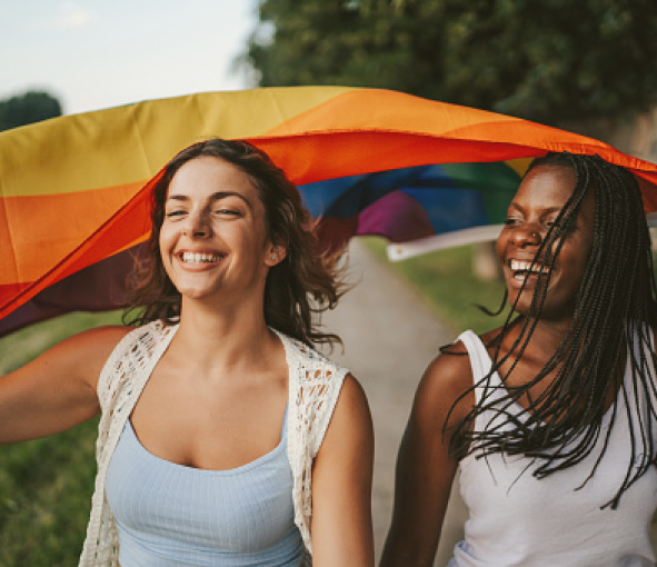 Two women smiling and carrying a rainbow flag billowing in the wind