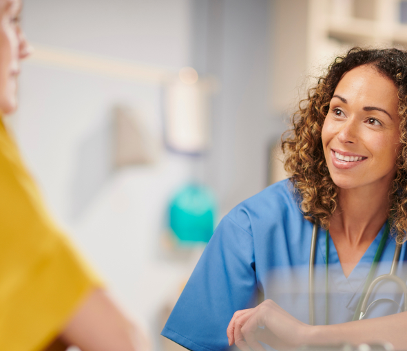 A woman in scrubs smiles while listening toΩ her patient